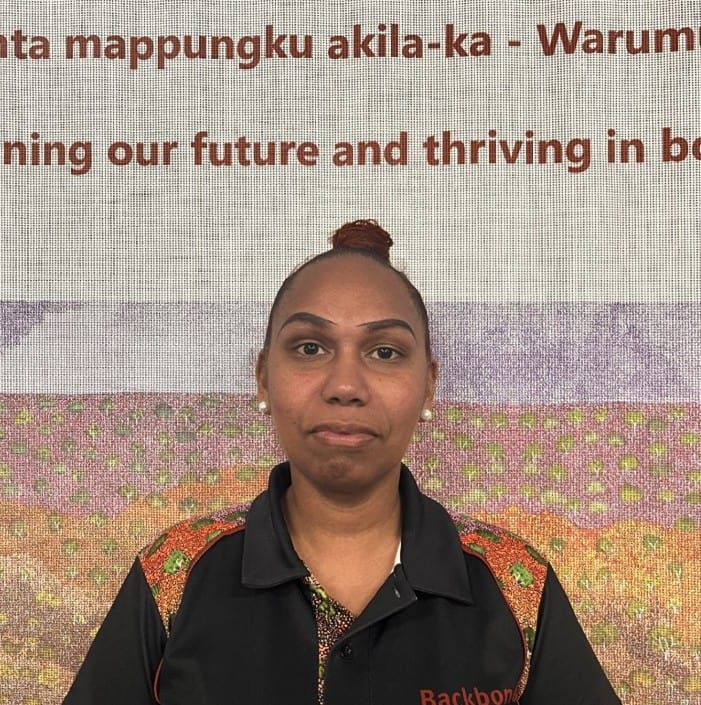 Portrait of a woman with a topknot bun, wearing a black polo with orange accents, standing in front of a screen with text.