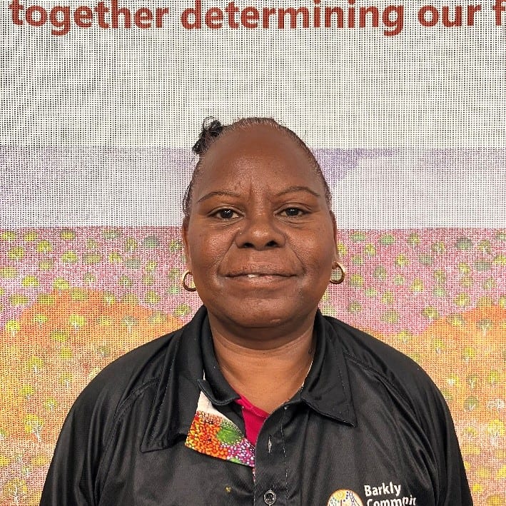 Portrait of a smiling Black woman with short hair, wearing a black jacket with a colorful patch, standing in front of a banner.