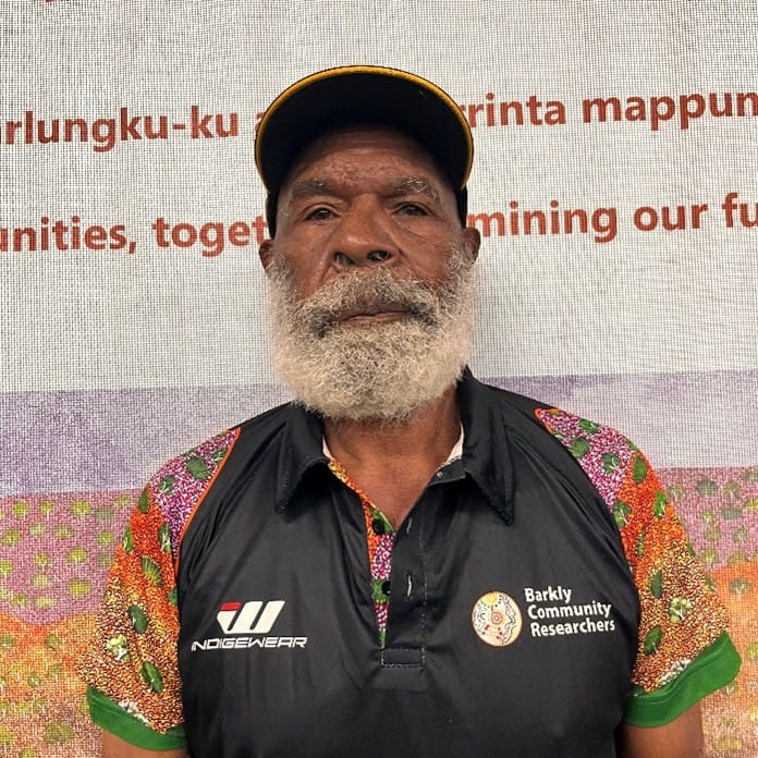 Portrait of an older man with a white beard wearing a yellow cap and a black polo with colorful sleeves, standing in front of a banner that reads 'Barkly Community Researchers'