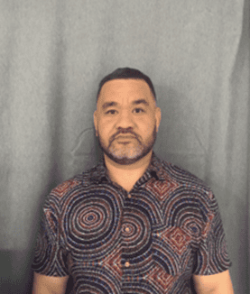 Man with short dark hair and beard, wearing a geometric-patterned shirt, standing against a gray curtain.