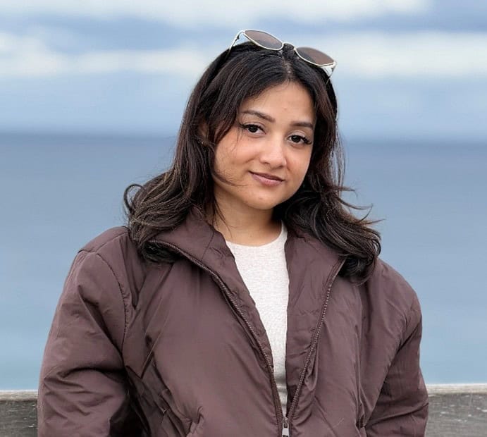 Young woman standing near a body of water, wearing a brown jacket and sunglasses on her head, looking at the camera.